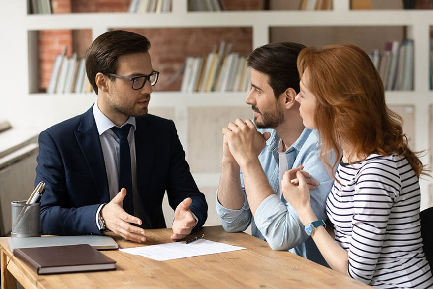 Meeting Discussing Three people sitting at a wooden table, reviewing a document, with one person in a suit gesturing while explaining.